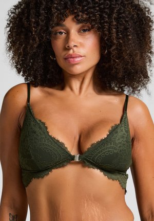 Vrouw met krullend haar die een olijfgroene kant bh met dunne bandjes en een sluiting aan de voorkant draagt, tegen een effen achtergrond.