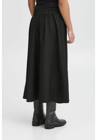 Schwarzer, langer Rock aus einem fließenden Material mit elastischem Bund. Das Model trägt kniehohe Stiefel. Glatte Textur, minimales Design.