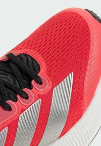 Red athletic shoe with a textured knit upper, black laces, and three silver diagonal stripes on the side; features a rounded white sole.