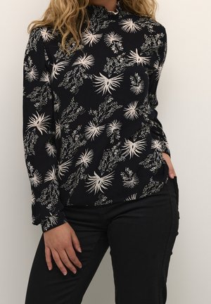 Frau trägt ein schwarzes Langarmoberteil mit weißen Blumenmustern und hohem Kragen, kombiniert mit schwarzen Hosen, steht mit einer Hand in der Tasche.