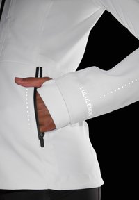 Veste de sport blanche avec fermeture éclair et poche, ornée du logo réfléchissant Lululemon et de accents en pointillés le long des manches. Design lisse et ajusté.