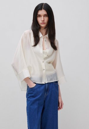 Frau mit langen dunklen Haaren, die eine lockere weiße Bluse mit Knöpfen über einem weißen Top und blaue hoch taillierte Jeans trägt, steht vor einem schlichten Hintergrund.