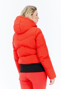 Mujer con chaqueta acolchada roja con capucha y pantalones rojos a juego, de perfil frente a un fondo blanco liso.