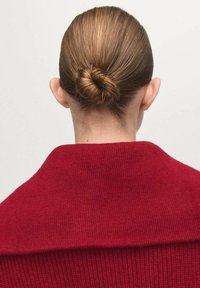 Roter Strickpullover mit hohem Kragen und geripptem Muster, getragen mit tief gebundenem Haar zu einem Dutt, zeigt eine glatte Oberfläche.