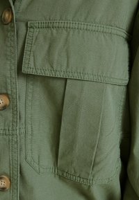 Grön skjorta med knäppning och bröstficka, mjukt tyg och sömdekoration. Knäppning med rundad knapdesign.