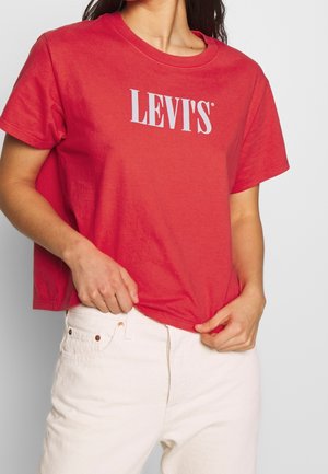 Kvinde iført en rød Levi’s t-shirt og hvide bukser, der holder i nederste kant af t-shirten med begge hænder.