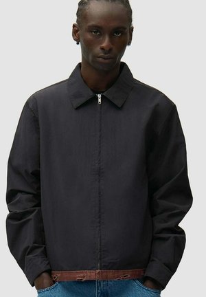 Uomo con capelli intrecciati che indossa una giacca nera con zip e orlo marrone consumato, mani in tasca, in piedi davanti a uno sfondo semplice.