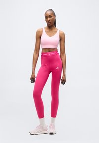 Γυναίκα με ροζ αθλητικό σουτιέν και κολάν, κρατώντας μαύρα αλτήρες βάρους 1 κιλού, στέκεται μπροστά σε καθαρό λευκό φόντο.