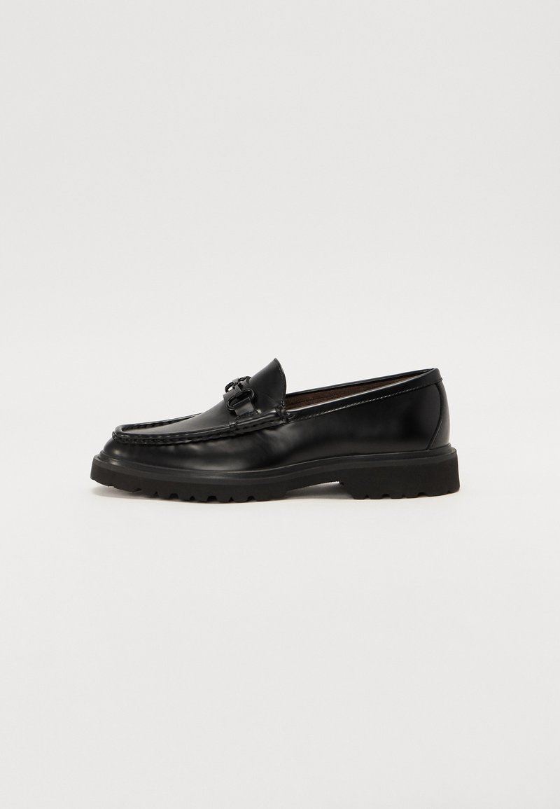 Mocassin en cuir noir avec un bout arrondi, une boucle décorative et une semelle en caoutchouc texturée avec des motifs de traction. Design classique avec des accents minimaux.