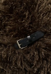 Ceinture en cuir noir avec une boucle argentée reposant sur un fond en tissu ou tapis en fourrure bouclée brun foncé.