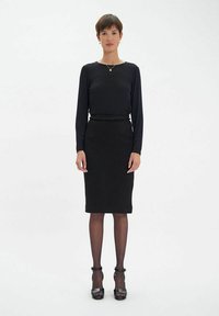 Haut noir à manches longues associé à une jupe noire à hauteur du genou. Il présente un détail texturé à la taille et des chaussures à talons hauts noires.