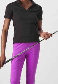 Person wearing a black short-sleeve shirt and bright purple checkered pants holding a golf club horizontally across their body.