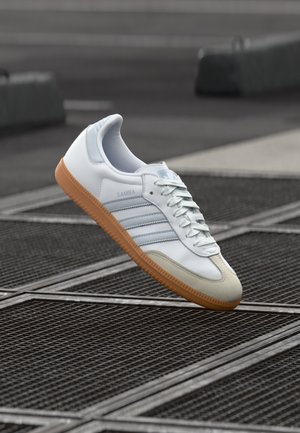 Weiße Adidas Samba Sneaker mit Gummisohle, hellblauen Streifen und beigem Zehenkappen, die über einer Metallgitteroberfläche im Freien schwebt.