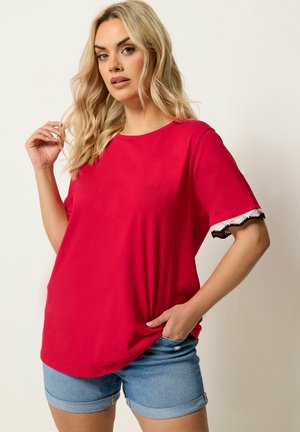 Blondynka w jasnoczerwonej koszulce z krótkim rękawem z falowanym wykończeniem na rękawach oraz w jasnoniebieskich podwiniętych szortach jeansowych.