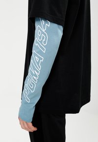 Schwarz T-Shirt mit lockerer Passform, das einen langen, hellblauen Ärmel hat, auf dem "PUMA 194" in Weiß entlang des Arms gedruckt ist.