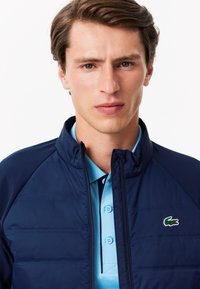 Giubbotto imbottito blu navy con collo alto, dotato di logo Lacoste verde. Indossato sopra una polo azzurra chiara con due bottoni e colletto.