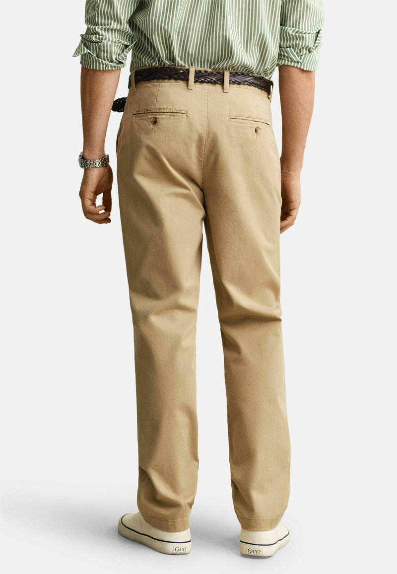 Mand iført beige bukser med baglommer, brunt flettet bælte, grønstribet skjorte, sølvur og hvide sneakers, der står med ryggen mod kameraet.
