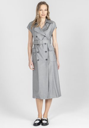 Kobieta ubrana w szarą, midi sukienkę bez rękawów z paskiem, szerokimi klapami i dużymi przednimi kieszeniami, w połączeniu z czarno-białymi loafersami.