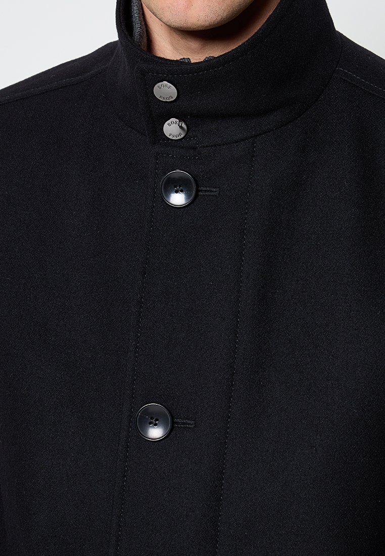 Manteau en mélange de laine noire avec un haut col, deux grands boutons et des coutures nettes. Texture douce avec des accents minimaux.