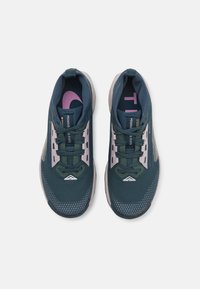 Zapatillas deportivas de color verde oscuro con acentos en gris claro, parte superior de malla, cuello acolchado y un logo rosa prominente en la lengüeta. Patrón hexagonal en la suela.
