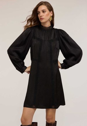 Robe noire avec des manches bouffantes longues, un col montant, des détails en œillets et un tissu texturé. Associée à des bottes montantes à motifs.