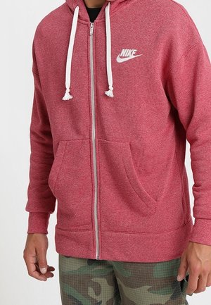 Uomo che indossa una felpa con cappuccio rossa Nike con cerniera e coulisse bianche, e pantaloni con motivo mimetico.