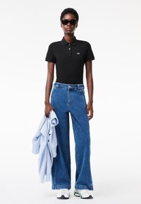 Svart pikétröja med korta ärmar, blå vida jeans i denim, ljusblå jacka i handen och vita sneakers med färgglada detaljer.