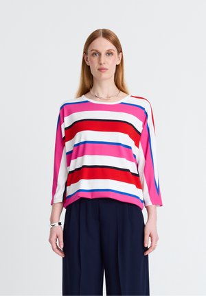 Frau mit glattem roten Haar, die ein lockeres gestreiftes Oberteil mit roten, pinken, blauen, schwarzen und weißen horizontalen Streifen und dunkle weite Hosen trägt.