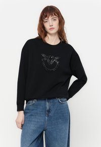 Črna sweatpanta iz mehkega blaga, z teksturirano zasnovo na sprednji strani in dekorativnimi akcenti. V kombinaciji s svetlo modrimi kavbojkami.