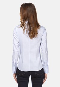 Chemise bleu clair à manches longues avec une coupe ajustée, poignets à boutons et texture lisse. Pas de motifs visibles, portée avec un jean sombre.