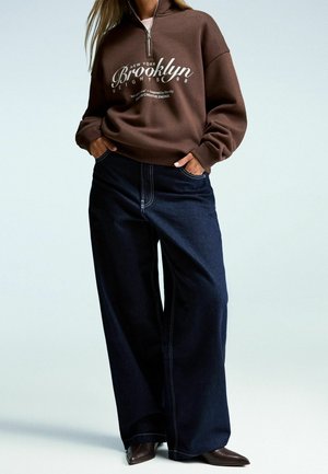 Kvinde iført brun quarter-zip sweatshirt med teksten "Brooklyn", mørke vidde bukser og brune spidse sko, står med hænderne i lommerne.