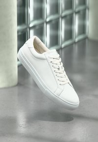 White leather sneaker with a smooth finish, cream laces, rubber sole, and embossed logo detail on the side, floating above a polished surface.