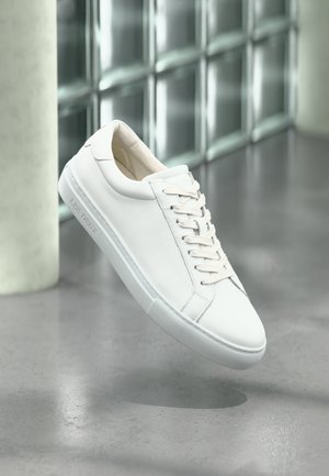 Baskets en cuir blanc avec une finition lisse, lacets crème, semelle en caoutchouc et détail de logo embossé sur le côté, flottant au-dessus d'une surface polie.