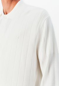 White knitted polo with vertical ribbing, featuring a collar and three buttons. Subtle logo detail in matching tone on the chest.