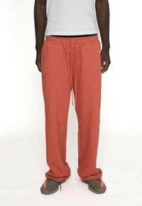 Mørk hudfarvet person iført løse rustfarvede sweatpants med snor og sorte-grå sneakers med orange detaljer, stående foran en hvid baggrund.