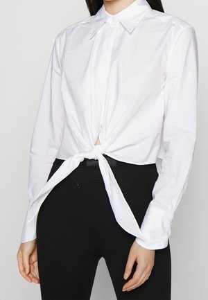 Chemise blanche à manches longues avec col noué devant au-dessus d’un pantalon noir moulant, portée par une personne aux longs cheveux foncés.