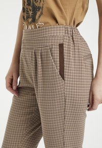 Bruna rutiga byxor med hög midja, veckad framsida och kontrasterande fickdetalj. Tillverkade av ett texturerat tyg.