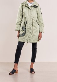 Cappotto lungo color verde chiaro, imbottito, con vita regolabile tramite coulisse, design trapuntato e dettagli in ricamo a forma di foglia nera; abbinato a jeans neri e tacchi aperti.