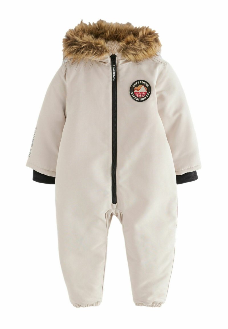 Combinaison isolante beige avec capuche doublée en fausse fourrure, fermeture éclair à l'avant, poignets élastiques et patch circulaire de la marque. Matériau doux et lisse.