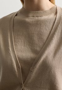 Beige kardigan vyrobený z měkké pleteniny, s výstřihem do V, zapínáním na knoflíky a žebrovaným límcem, navrstvený přes odpovídající top.
