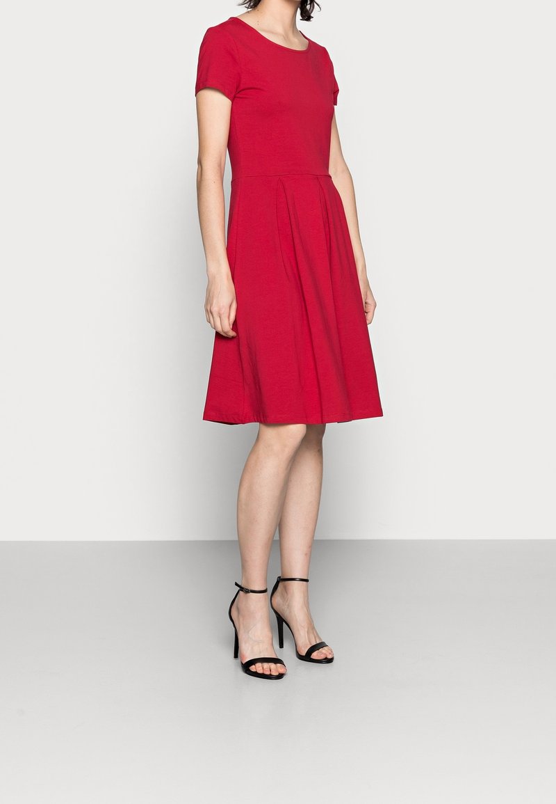 Punainen lyhythihainen mekko, joka on valmistettu puuvillasta, vyötäröllä on laskoksia, polvipituinen helma, ja se on yhdistetty mustiin naruilla varustettuihin korkokenkiin.