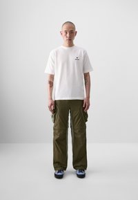 T-shirt en coton blanc avec un petit logo noir sur la poitrine, associé à un pantalon cargo vert olive avec plusieurs poches et un fit décontracté.