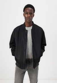 Chaqueta bomber negra con cierre frontal, cuello acanalado y mangas largas, llevada sobre un suéter acanalado gris claro, combinada con jeans grises.