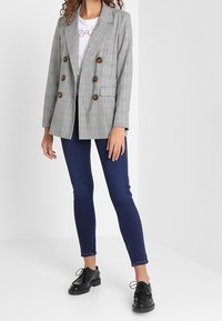 Blazer de cuadros gris de doble botonadura con botones marrones, llevado sobre una camisa blanca y combinado con jeans ajustados azul oscuro y zapatos negros con cordones.