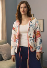Giacca trapuntata floreale bianca con vivaci motivi blu e rosa, rifinita con un bordo e delle stringhe corallo a contrasto. Abbinata a una canotta bianca.