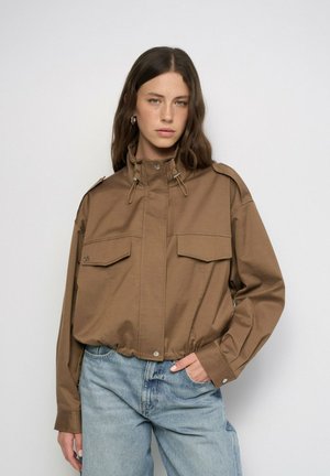 Mujer con cabello largo y castaño que lleva una chaqueta marrón corta con grandes bolsillos y vaqueros azul claro, de pie frente a un fondo liso.
