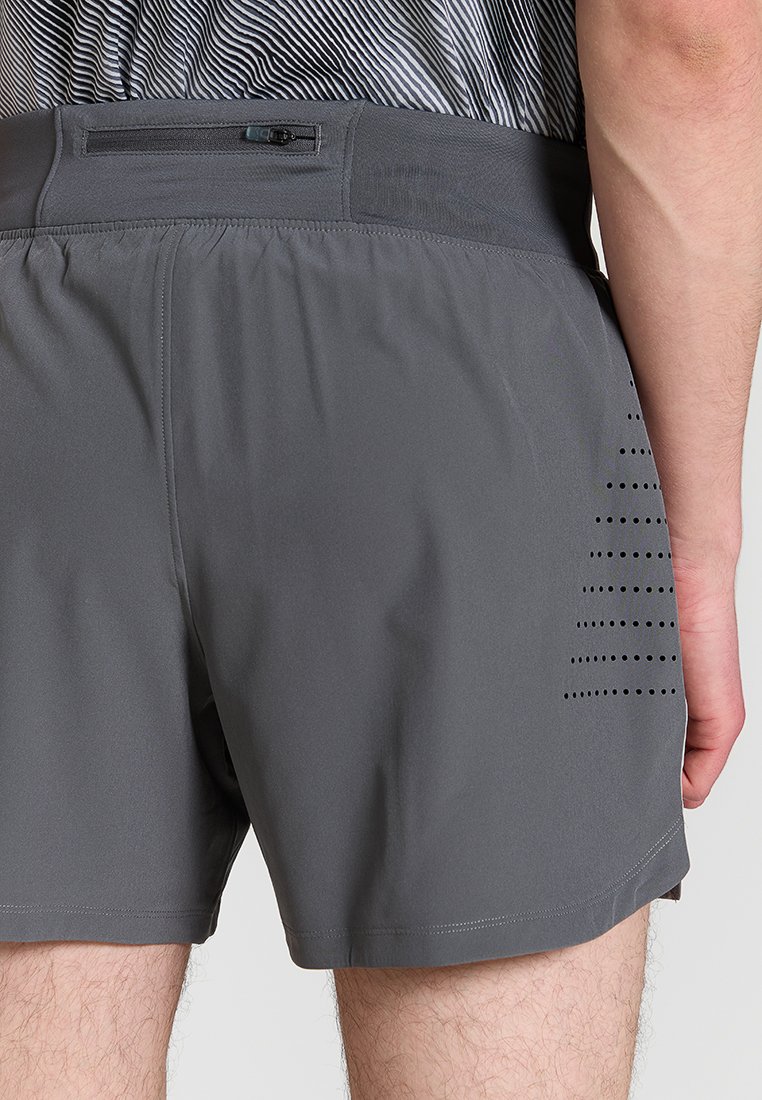 Uomo che indossa pantaloni sportivi grigi con tasca posteriore con zip e ventilazione forata sul lato destro, camicia a righe infilata nei pantaloni.