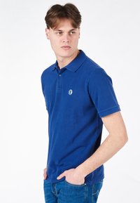 Polo blu realizzato in cotone lavorato a maglia, con colletto, maniche corte e un piccolo logo sul lato sinistro del petto. Abbinato a jeans in denim.