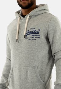 Sudadera gris claro con un bolsillo canguro frontal, cordones y texto bordado en azul. Material de algodón suave con un diseño casual.