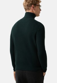 Mann mit Brille, der einen dunkelgrünen, gerippten Rollkragenpullover trägt, steht mit den Händen in den Taschen und blickt mit dem Rücken zu einer schlichten Wand.